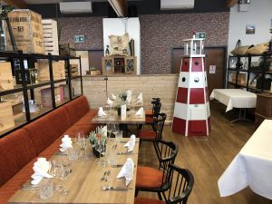 Restaurantbereich mit eingedeckten Holztischen, orangefarbener Sitzbank und einem rot-weißen Dekorationsleuchtturm.