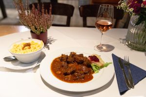 Ein Teller Gulasch mit einer Birne und Preiselbeeren, daneben eine Schüssel Spätzle und ein Glas Roséwein.