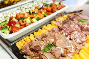 Buffetplatten mit Parmaschinken, Salami und Melone neben Schalen mit buntem Bauernsalat und Fetakäse.