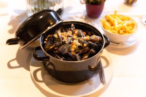Ein schwarzer Topf mit gekochten Miesmuscheln in einer hellen Soße, daneben eine Schale Pommes Frites.