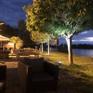 Beleuchtete Outdoor-Lounge am Abend mit Rattan-Sesseln und angestrahlten Bäumen am Gewässer.