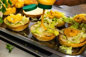 Eine Hand gibt Soße aus einer Flasche auf aufgeschnittene Burgerbrötchen mit Salat.