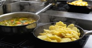 Zwei dampfende Pfannen auf einem Herd, eine mit Gemüseeintopf und die andere mit Bratkartoffeln.