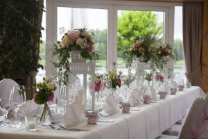 Festlich eingedeckte lange Tafel mit weißen Laternen, Blumengestecken und Gläsern vor einer großen Fensterfront.