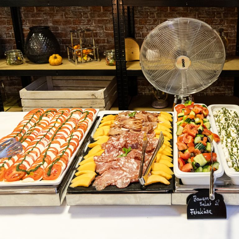 Kaltes Vorspeisenbuffet mit Tomate-Mozzarella, Schinken mit Melone, Bauernsalat, mariniertem Fetakäse und Antipasti vor einem Tischventilator.