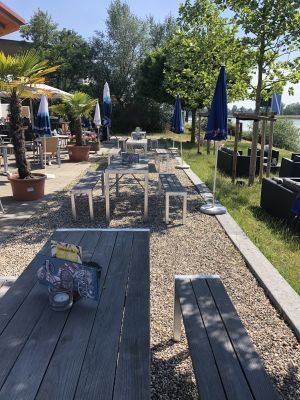 Sitzbereich auf Kies mit grauen Holzbänken, blauen Sonnenschirmen und Kübelpalmen am Ufer.