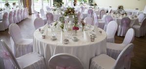 Festlich eingedeckte runde Tische mit weißen Stuhlhussen, rosa Schleifen und Blumenschmuck in einem Veranstaltungssaal