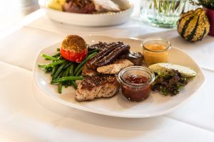 Ein weißer, ovaler Teller mit gegrilltem Fleisch, Würstchen, grünen Bohnen, einer gefüllten Tomate und Dips.