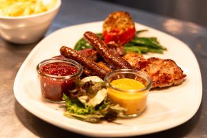 Ein Grillteller mit Würstchen, Fleisch, grünen Bohnen, gefüllter Tomate und Dips.