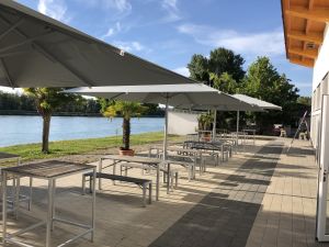 Gepflasterte Außenterrasse am Gewässer mit großen Sonnenschirmen, Stehtischen und Kübelpalmen.