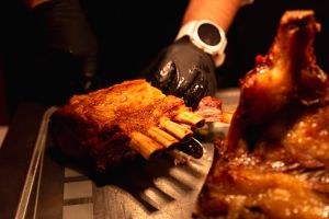 Hände mit schwarzen Handschuhen schneiden einen großen Braten, ein Lammkarree, auf einem Metallblech.