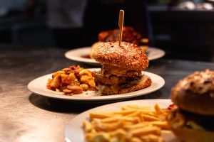 Ein hoher Burger mit Sesambrötchen auf einem Teller, daneben eine Portion Bratkartoffeln mit Speck.