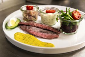 Ein weißer Teller mit Matjesfilets, Soßen, einem Beilagensalat im Glas und einem Stück Ei.