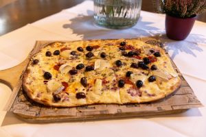 Ein rechteckiger Flammkuchen auf einem Holzbrett, belegt mit Käse, schwarzen Oliven und roten Zwiebeln.