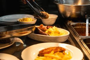 Eine Zange legt eine Wurst in eine Schüssel mit Kartoffelpüree und Fleisch in einer Restaurantküche.