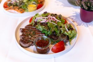 Ein weißer Teller mit aufgeschnittenen Rindersteakstreifen, einem Beilagensalat und einem Glas Dip.